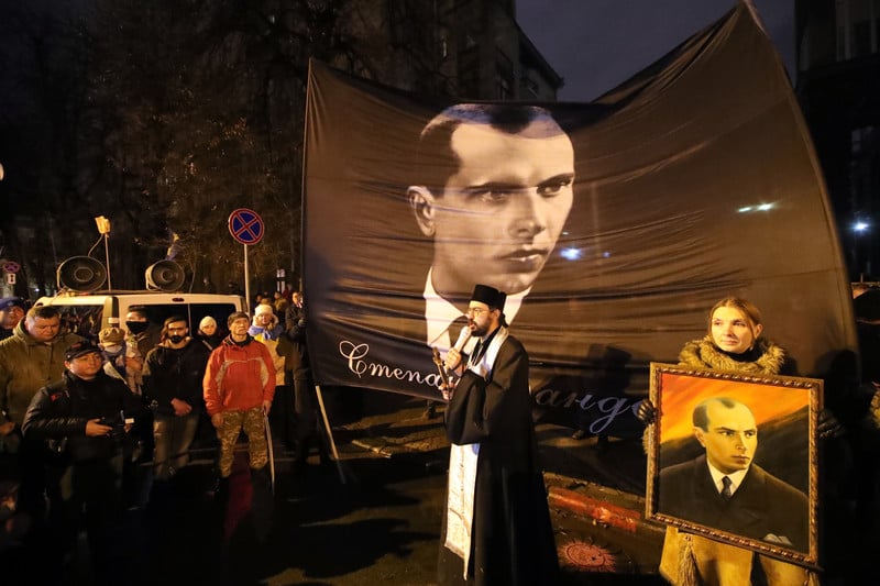 People watch a man in religious garb speak in front of a huge portrait of Stepan Bandera