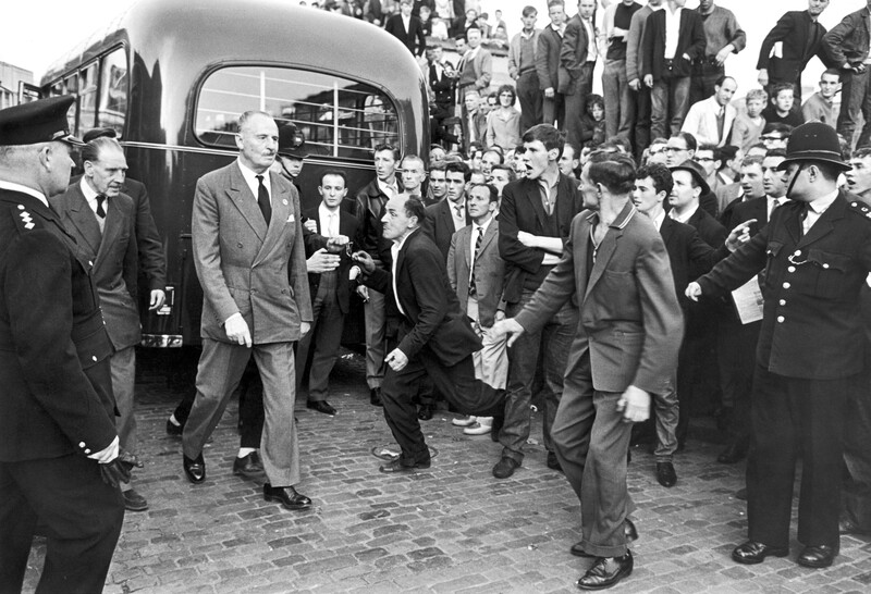 A man in a suit walks through an angry crowd not quite held back by uniformed police officers