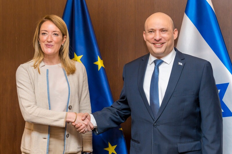 Woman and man shake hands and smile in front of EU and Israeli flags.