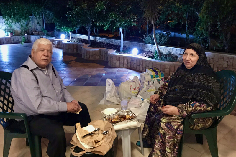 Man and woman sit on chairs on a paved area surrounded by trees in the evening