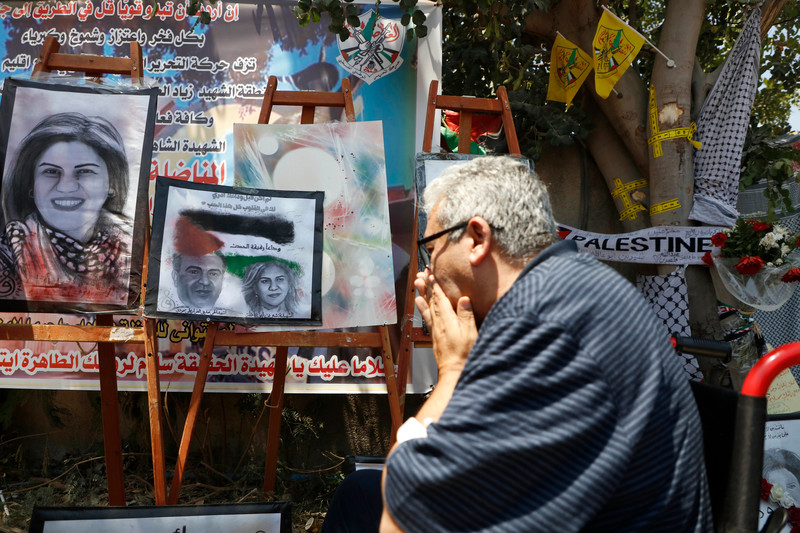 A man holds his hands to his face as he passes by posters of slain correspondent Shireen Abu Akleh