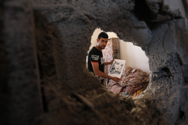 A man holds a drawing through a hole in a wall caused by the Israeli bombing campaign in early August