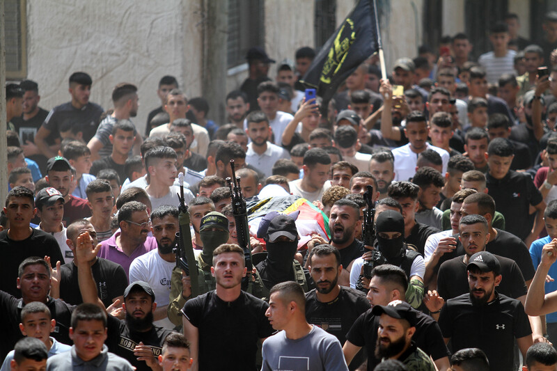 Men carry a body on a stretcher in a crowd that includes some with rifles 