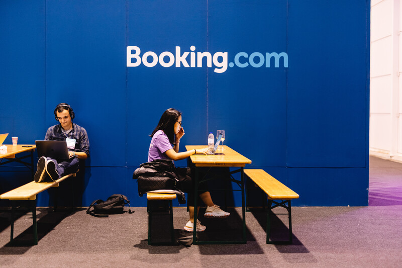 Two people sit on benches and work in front of a Booking.com wall