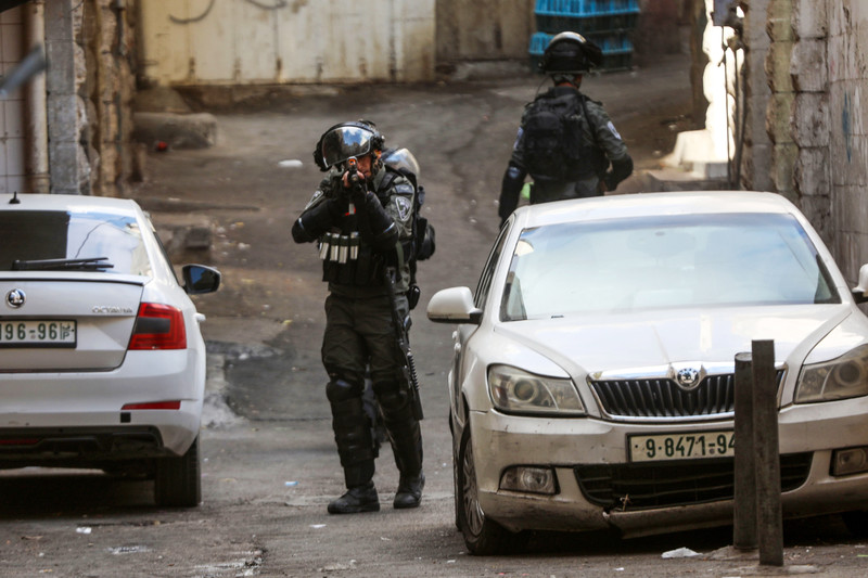 An Israeli Border Police combatant aims a weapon towards camera while standing between cars on street