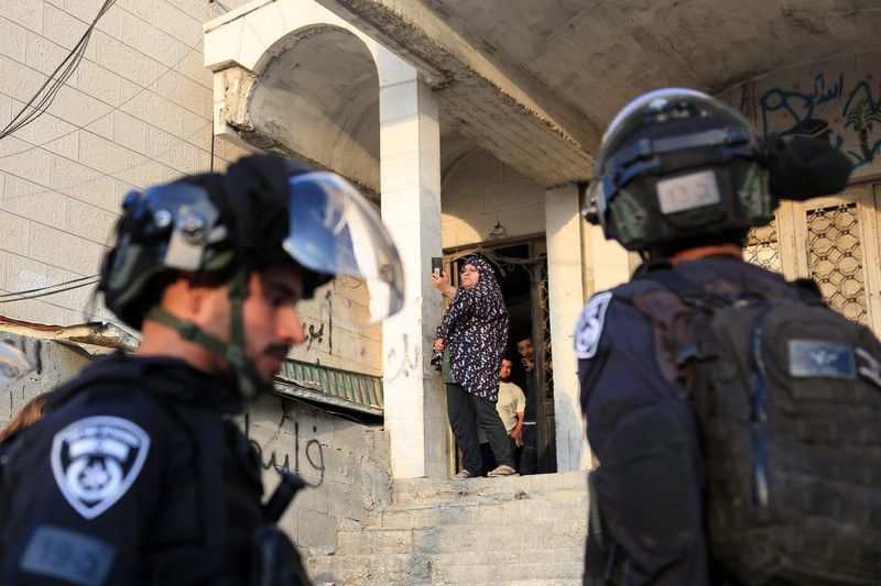 Palestinian woman stands atop staircase in front of home with Israeli police in foreground
