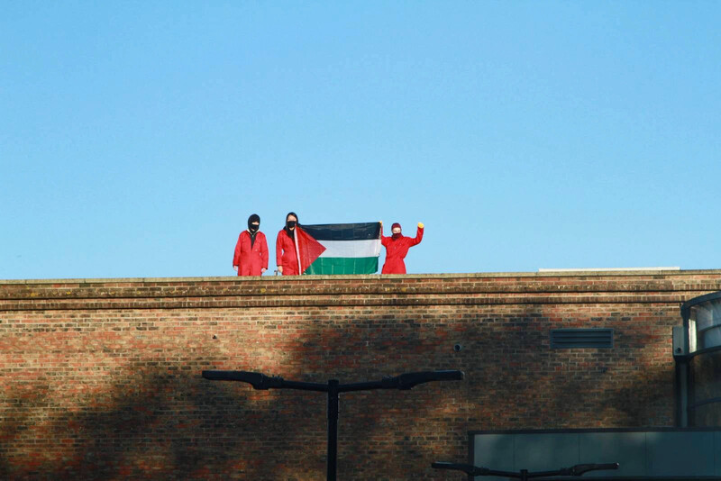 Three people in red hold up a Palestinian flag on a rooftop