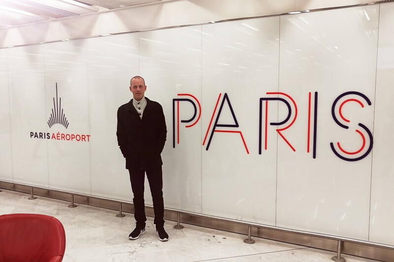 A man stands next to a sign reading Paris Airport and Paris