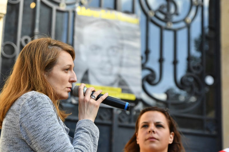 A woman holds a microphone in front of poster of a man as another woman looks on