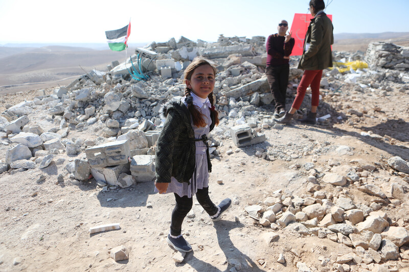 Girl in school uniform stands in front of rubble with Palestinian flag flying over it as two other people stand behind