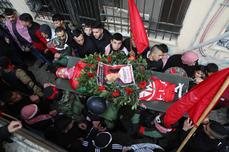 Men and boys carry the body of a boy wrapped in a flag and covered in a wreath and a poster on a stretcher 