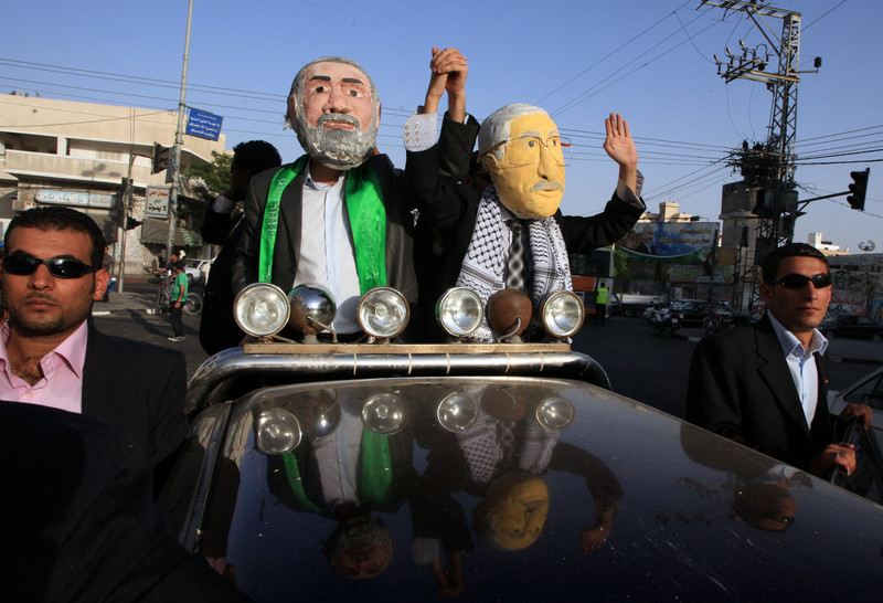 Two mean wearing masks depicting Ismail Haniyeh and Mahmoud Abbas wave from a black car