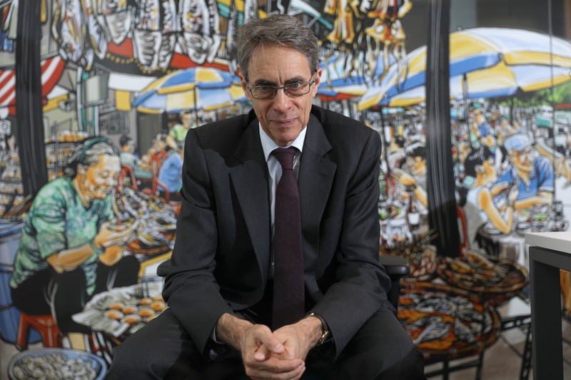 A man in a suit sits in front of a mural of a lively street scene
