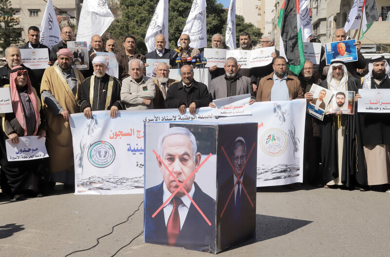 People hold signs, flags and banners in front of a cube structure that has the faces of Israeli government officials crossed out 