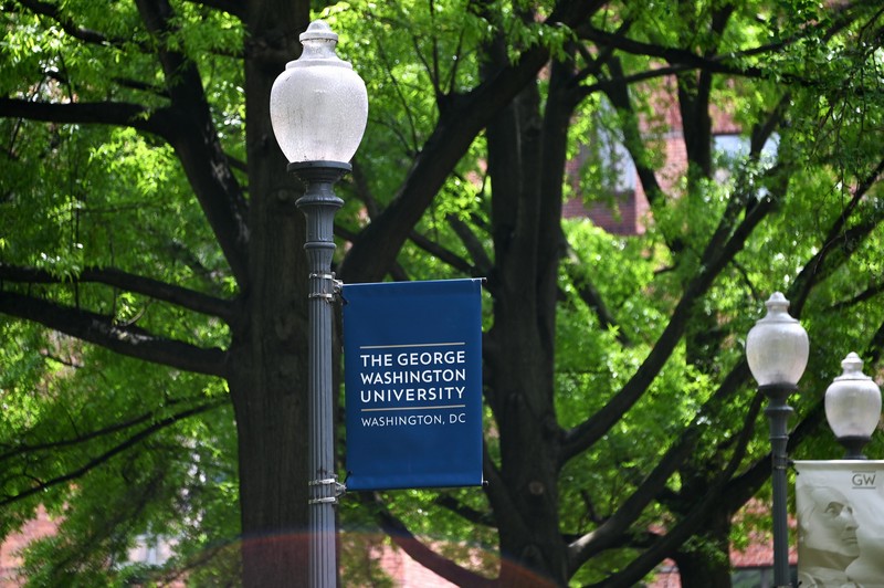 A blue banner with the name of the university hangs on a black lamppost, with trees in the background 