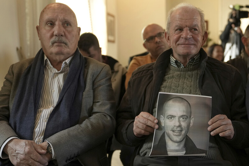 Two elder men sit next to one another and one holds a photo of a younger man