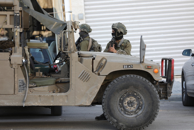 Two soldiers stand near armored vehicle, one of them pointing a sniper 