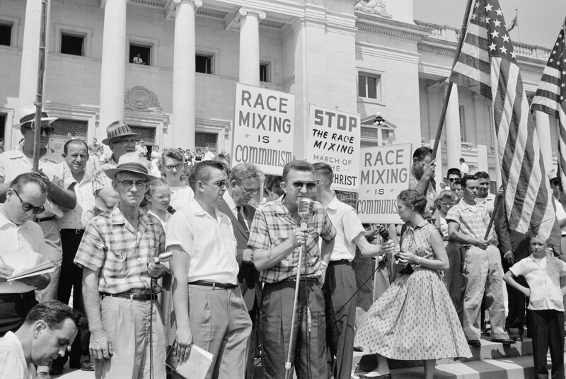 Man speaking at microphone in front of crowd at the Arkansas State Capitol protesting school integration with signs reading Race mixing is Communism and Stop the race mixing