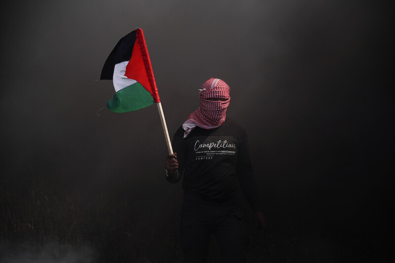 A person with their face covered emerges through dark smoke with the Palestinian flag