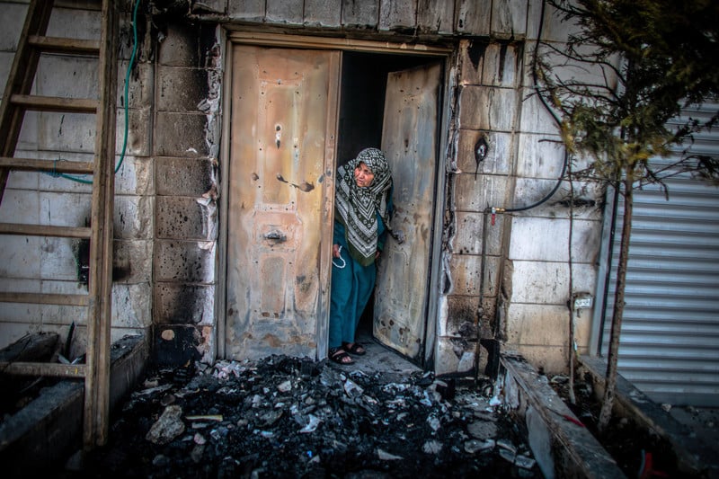 A women peers out from a metal door to an area covered in burned debris