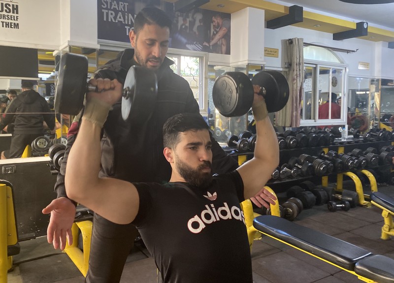 In a gym, one man spots for another man as he lifts two dumbbells.