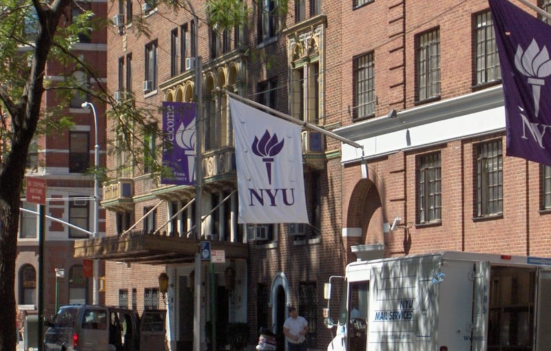 Banners with the NYU logo hang from brick buildings. A US mail truck is parked on the side of the street.