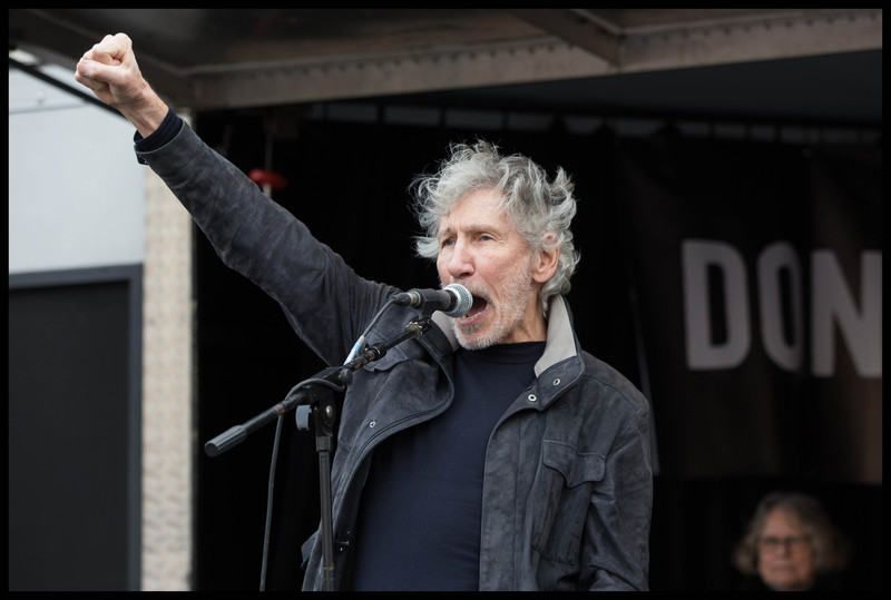 Roger Waters, dressed in a black t-shirt and a black jacket, stands at a microphone with his fist in the air
