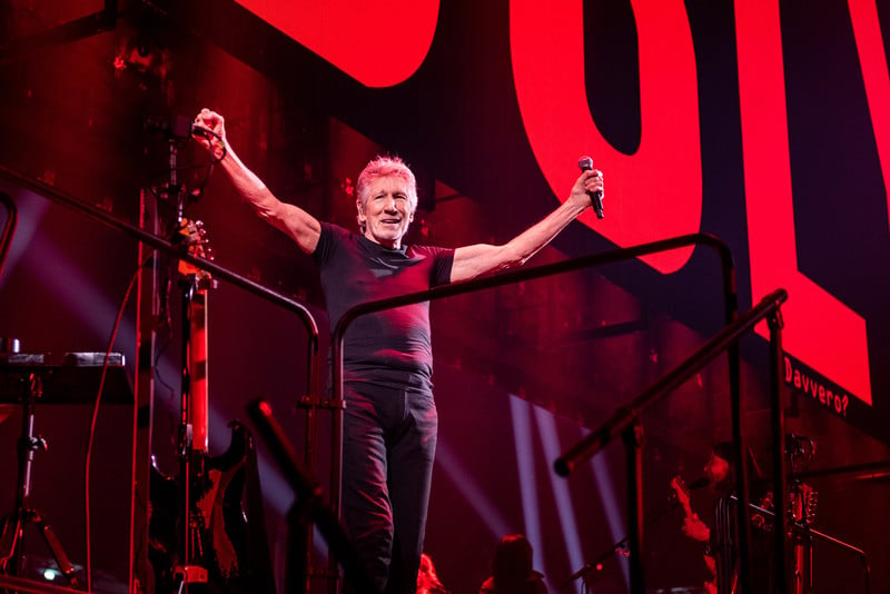 Roger Waters, dressed in a black t-shirt and black jeans and holding a microphone, raises his arms and smiles onstage