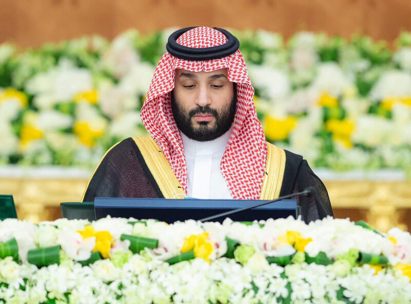 A man wearing a head dress and a gold-trimmed cloak looks down towards a podium surrounded by flowers