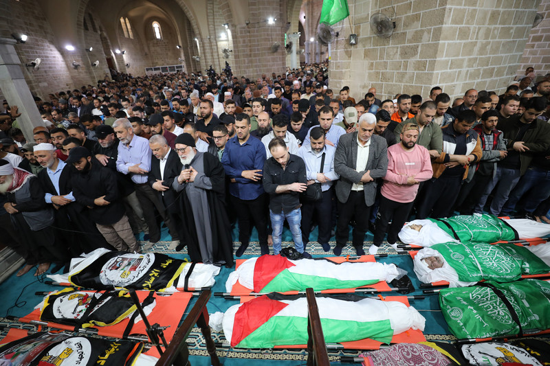 Men stand while praying while facing bodies wrapped in Palestine and faction flags on ground in front of them inside mosque