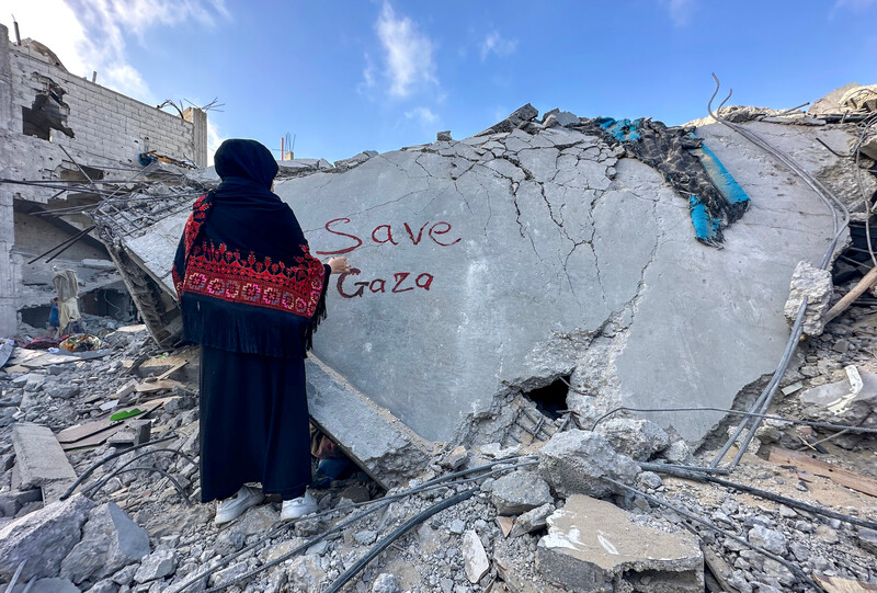 A young woman stands amid rubble of a home with the words Save Gaza written in red on a piece of concrete