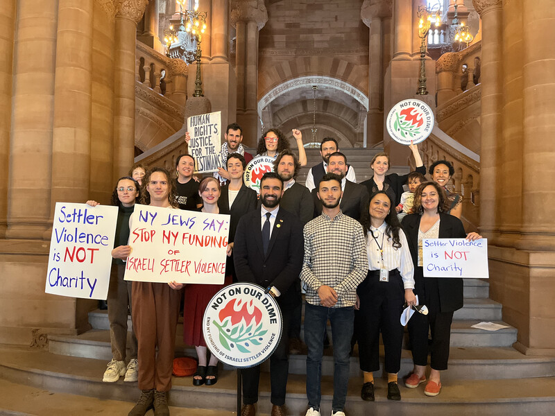 A group of people hold signs and banners supporting the Not On Our Dime campaign. Lawmaker Zohran Mamdani is at center, wearing a black suit.