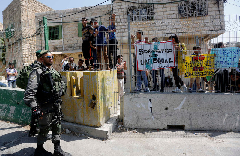 An Israeli soldier stands in front of a group of children who are behind a fence on a street. They are holding signs that denounce apartheid and segregation.