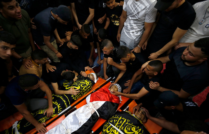 Men and boys sit next to and lean over three bodies on stretchers shrouded in Islamic Jihad and Palestineflags