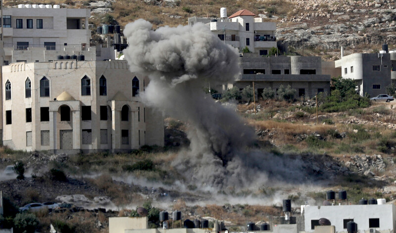 A plume of gray smoke rises over a field surrounded by buildings