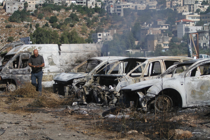 Man looks at phone in his hand while walking past the skeletons of burned-out cars with residential homes on hilly landscape in background