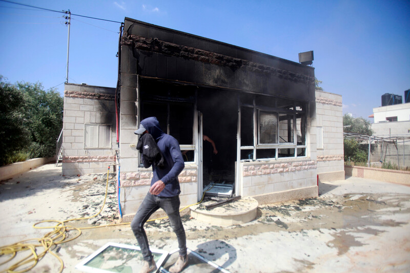 Man holds cloth to his face while walking in front of scorched single-story residential building with its front door and windows destroyed