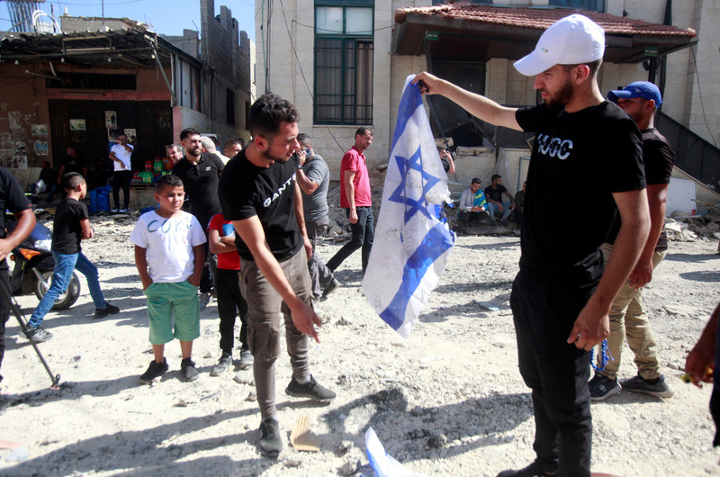 Man holds up Israeli flag while another man points to a flag on the ground as people stand and walk around rubble-strewn street