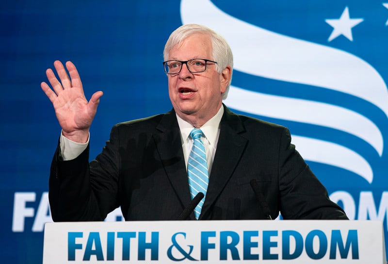 Dennis Prager raises his hand at podium before a faith and freedom sign