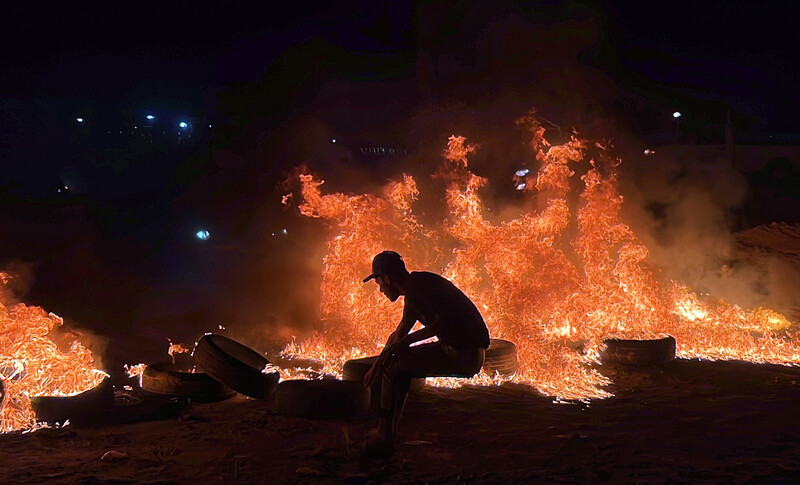 The silhouette of a man wearing a cap against the backdrop of fire and burning tires 