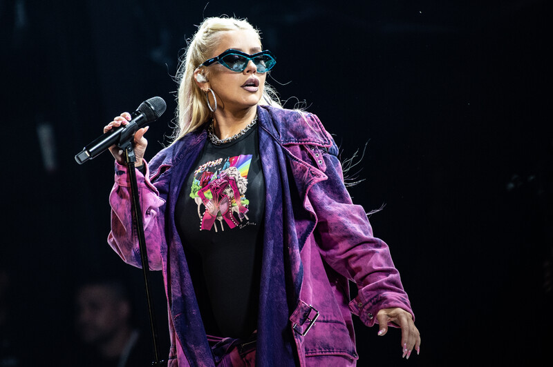 Christina Aguilera, in a purple jacket and wearing sunglasses, holds a microphone