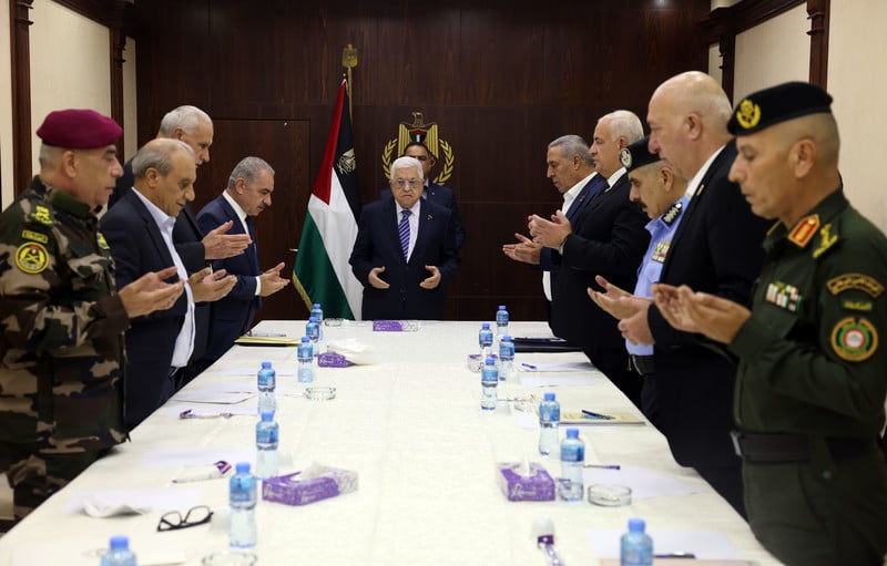 Men, some in uniform and others in suits, stand around a meeting table with Abbas at its head