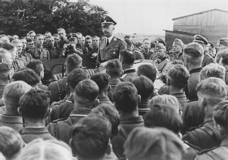 An older man in round spectacles and Nazi uniform stands in a car surrounded by young men in Nazi uniforms