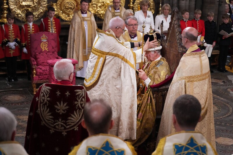 Welby in gold-trimmed bishop's robes places ornate crown on head of seated, robed monarch Charles as others look on