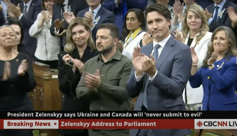 Zelensky in a military shirt stands next to Trudeau who is wearing a suit and clapping as others look on
