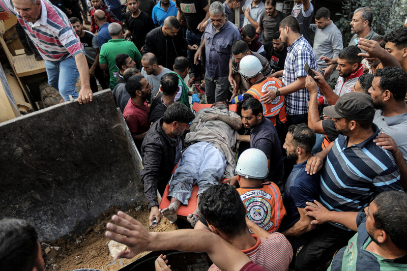 A crowd of men including rescue workers carry a man on a stretcher next to the scoop of an excavator machine