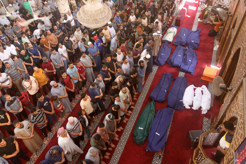 Men and boys stand with their arms crossed in prayer with the shrouded bodies of 13 people, three of them small children, inside a mosque