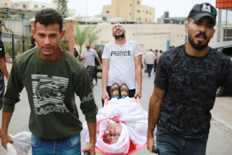 A man winces his face while carrying a bloodied person wrapped in a white sheet on a stretcher