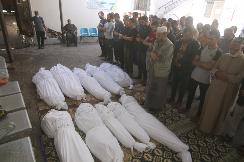 Men cross their arms in front of their stomachs and look down while standing in front of shrouded bodies of 10 people