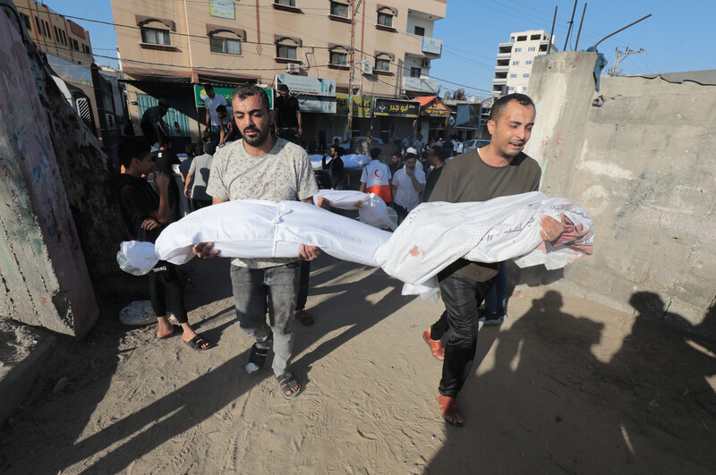 Two men carrie shrouded bodies while walking on sandy road with more people carrying bodies further behind them